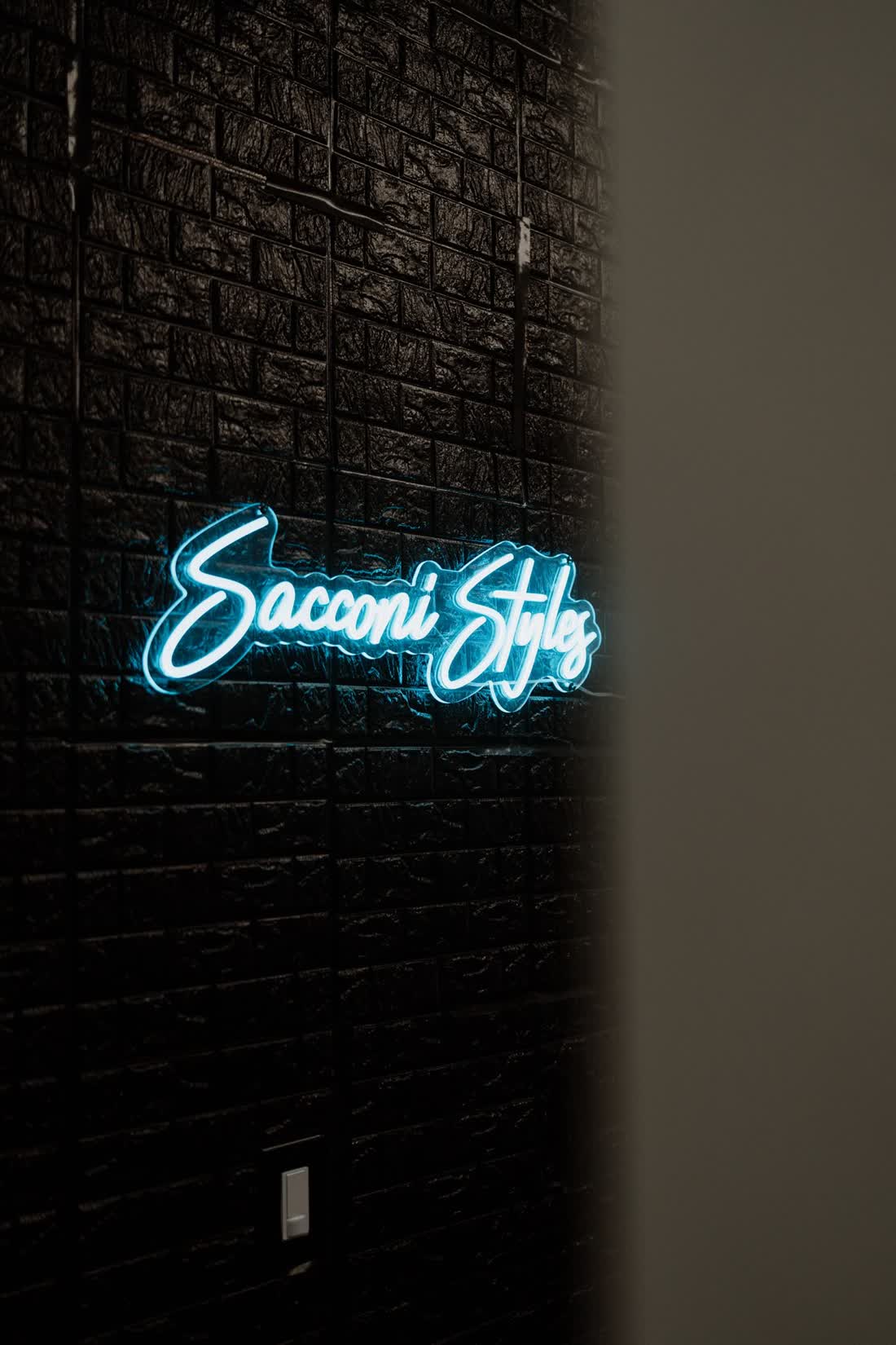 The glowing Sacconi Styles script sign on the black brick wall of Suite 102